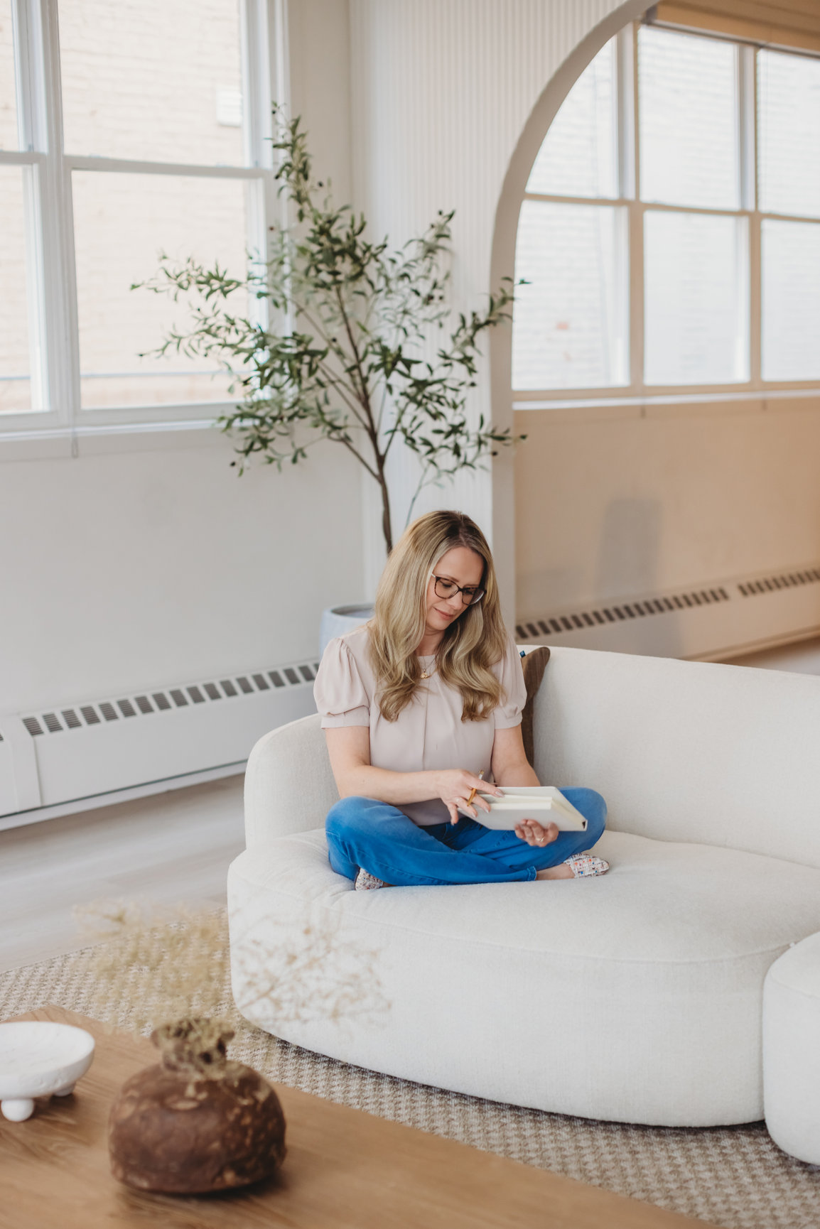 Sarah Hope sitting cross-legged on a cream bouclé sofa, journaling in bright window light